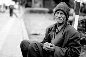A black and white portrait of an elderly man sitting on the streets, expressing hardship and resilience.